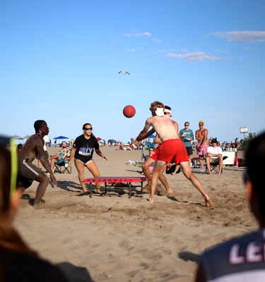 Volleyball players playing at a boardball sport tournament on the beach in Toronto, during the summertime. Spectators watch on the sidelines.